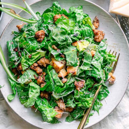 a vegan caesar salad with crumbled crispy tempeh and croutons on a white table with a gold fork