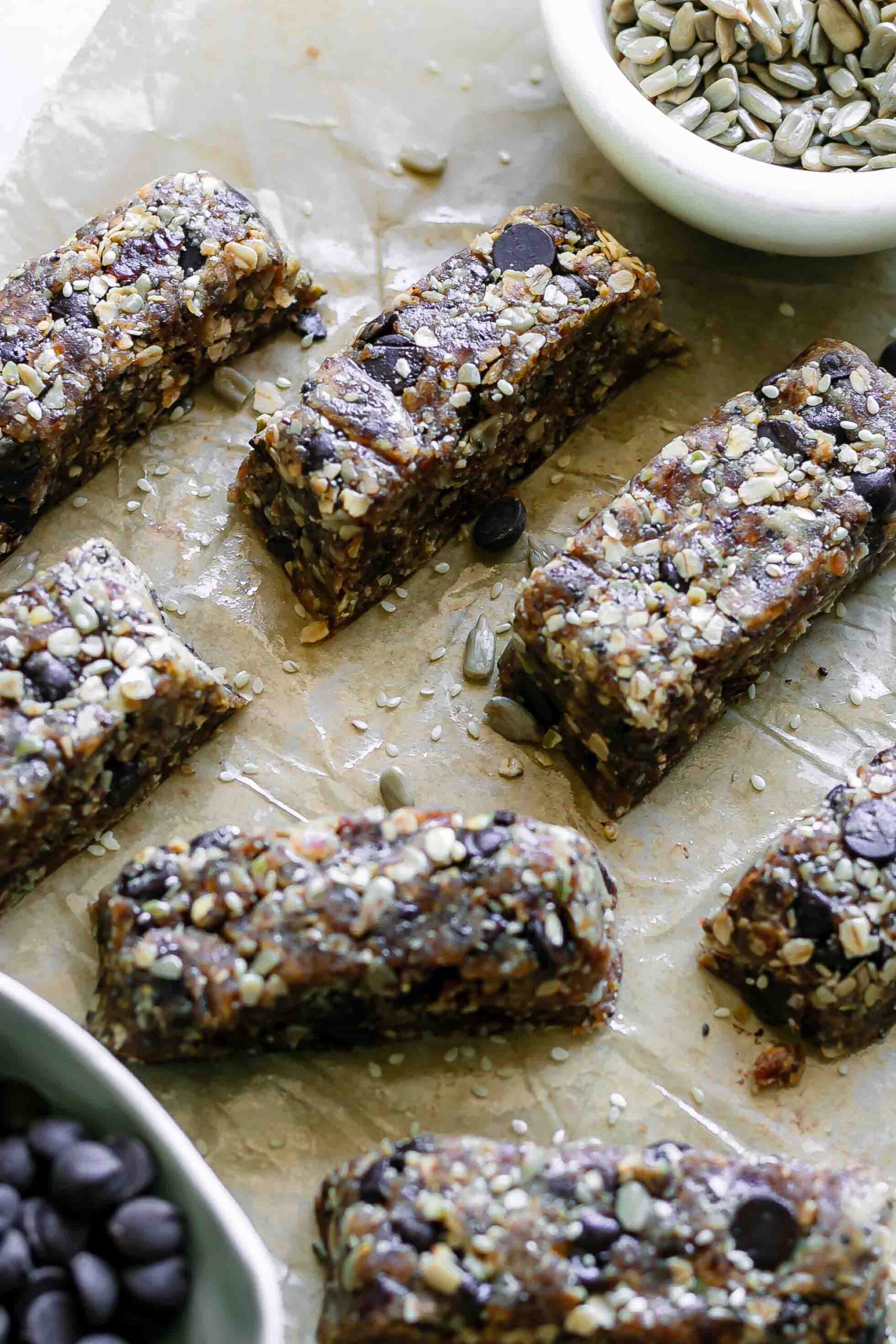 tahini oat and seed snack bars with chocolate on a brown table with bowls of seeds and chocolate chips