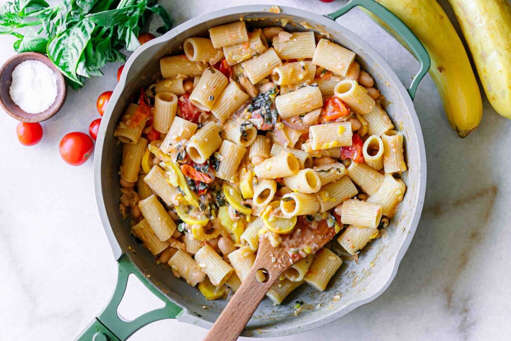 pasta in a creamy white bean sauce with roasted squash, cherry tomatoes, onions, and basil on a white table with a wooden spoon