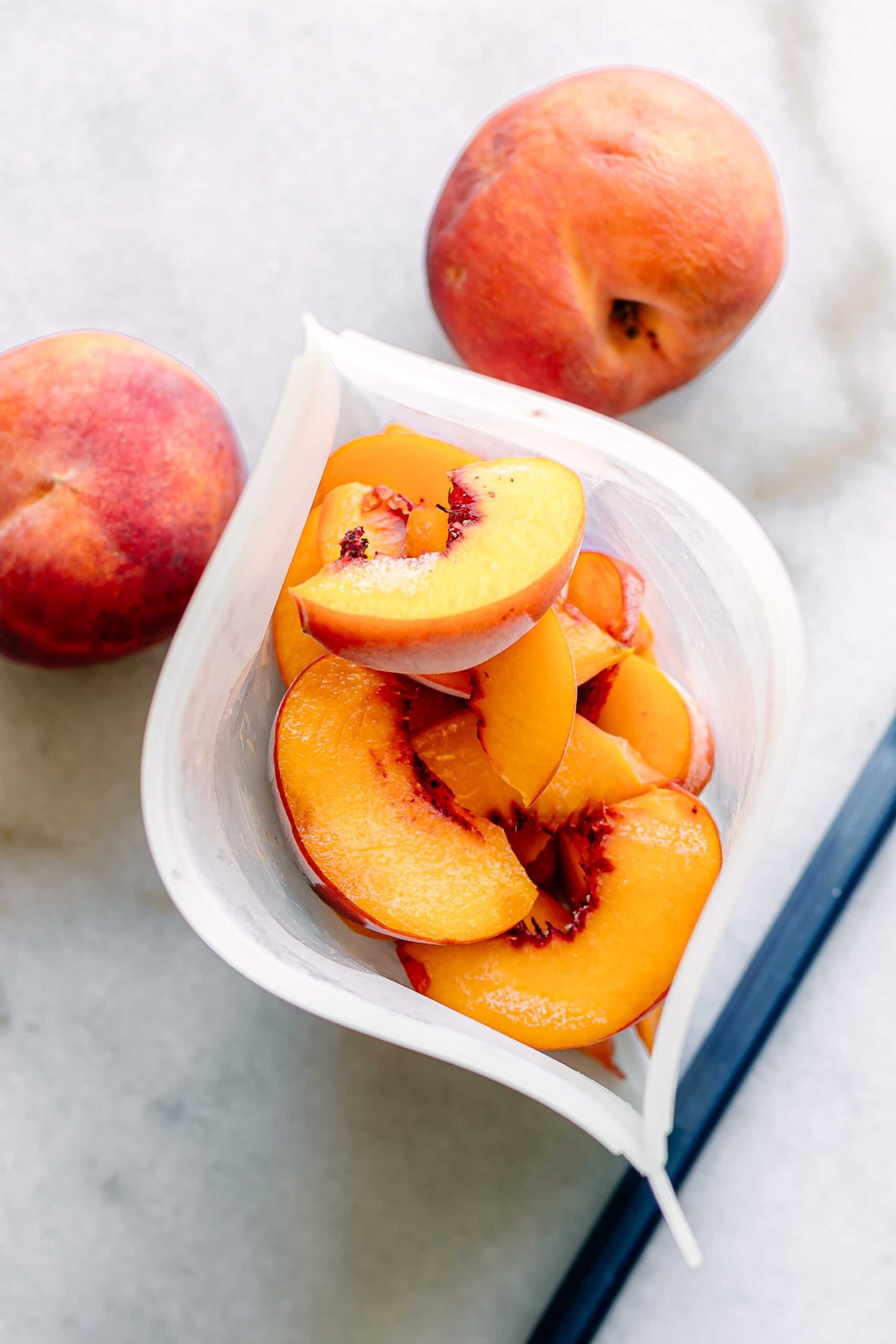 frozen peach slices in a silicone bag on a white table with two fresh peaches