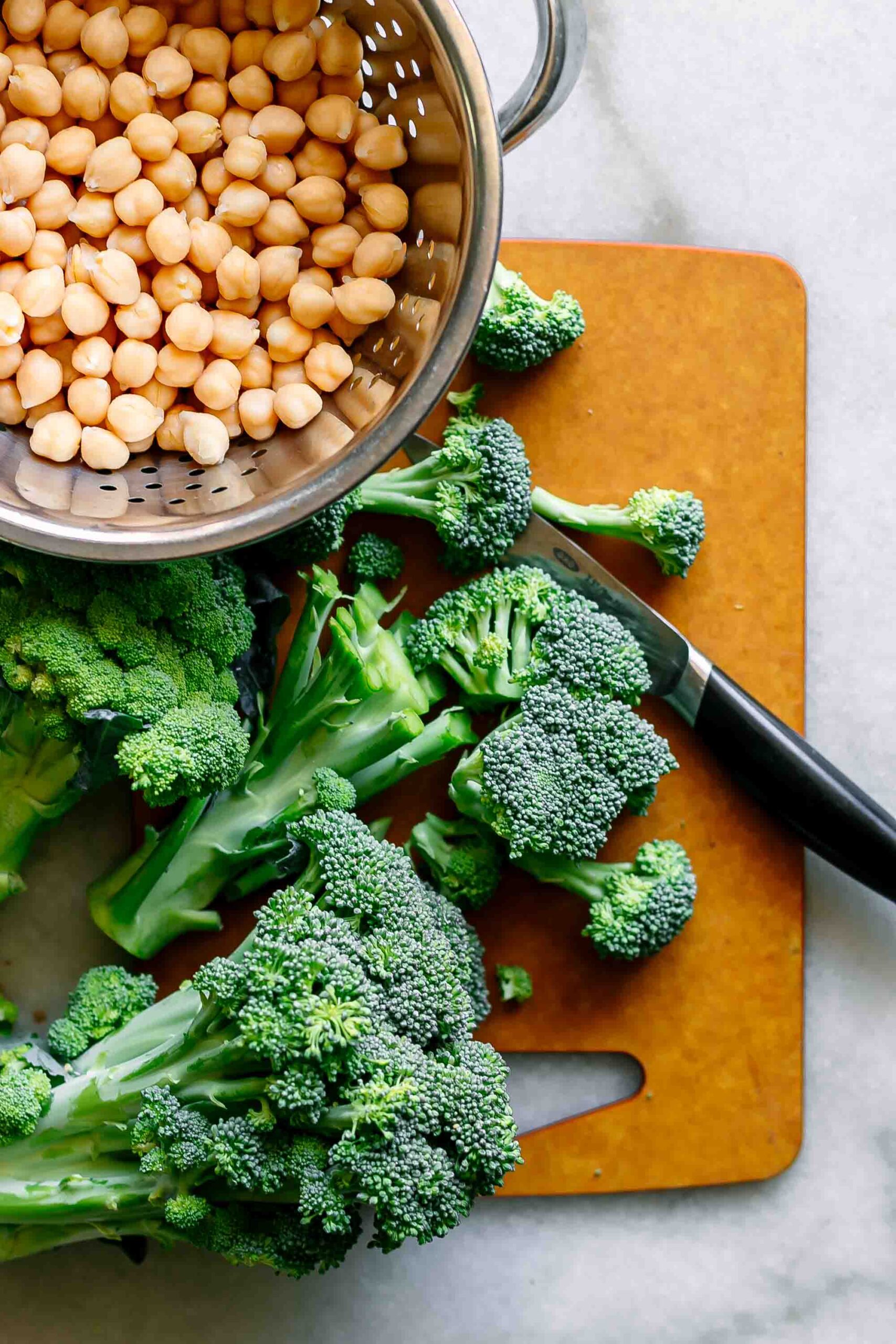 cut broccoli on a cutting board with a bowl of strained and washed chickpeas