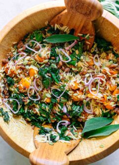 a large wooden bowl with an orzo and kale salad with butternut squash on a white table