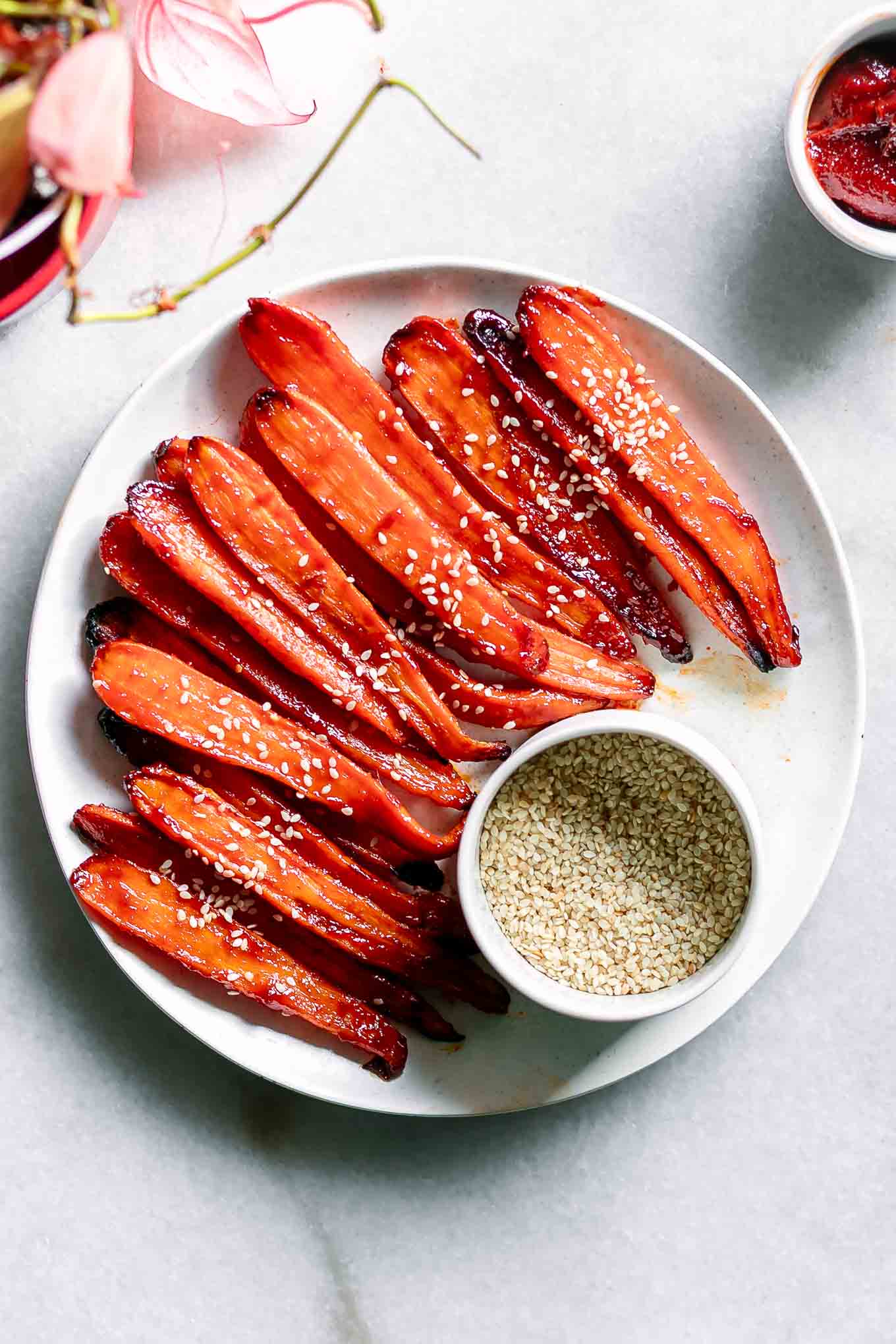 gochujang carrots on a white plate with a bowl of sesame seeds on a white table