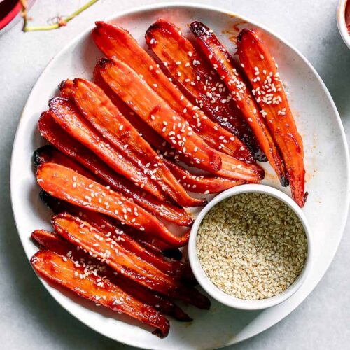 a plate with gochujang glazed carrots on a white table and a bowl of sesame seeds